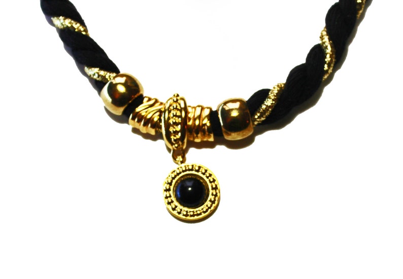 May include: A black and gold necklace with a braided cord. The necklace features gold-colored beads and a central pendant with a black stone. The pendant is surrounded by a gold-colored frame.