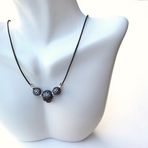 Op de afbeelding: Een zwarte koordketting met drie schedelvormige hangers. De centrale schedel heeft een rode edelsteen als oog, en de andere twee hangers zijn rond. De hangers zijn bedekt met kleine, glinsterende zwarte stenen.