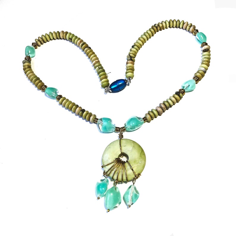 May include: A handcrafted necklace featuring a light green circular pendant with wire detailing and dangling aqua-colored teardrop-shaped beads. The necklace includes light green and tan-colored beads, aqua-colored accent beads, and a blue clasp.
