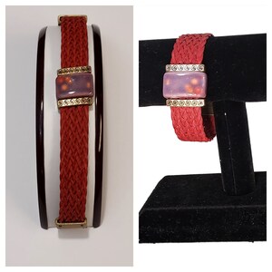 May include: A red braided bracelet with a rectangular purple and gold centerpiece. The bracelet is displayed on a black stand. The centerpiece has a gold border and a purple background with orange and yellow speckles.