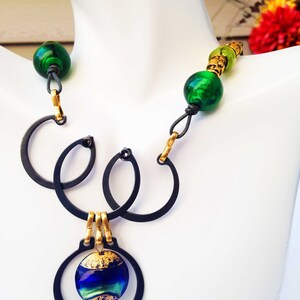 May include: A unique necklace featuring a combination of black metal hoops, gold accents, and vibrant green and blue glass beads. The pendant showcases a circular blue and gold glass element, adding a touch of elegance to the design.