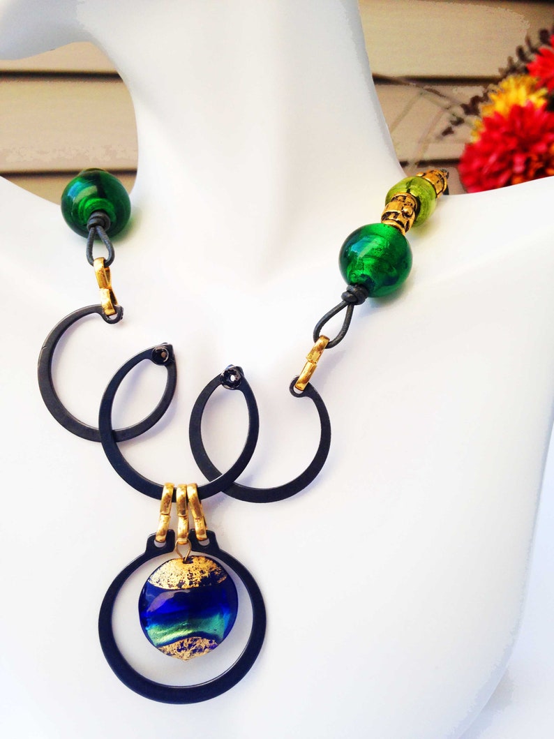 May include: A unique necklace featuring a combination of black metal hoops, gold accents, and vibrant green and blue glass beads. The pendant showcases a circular blue and gold glass element, adding a touch of elegance to the design.