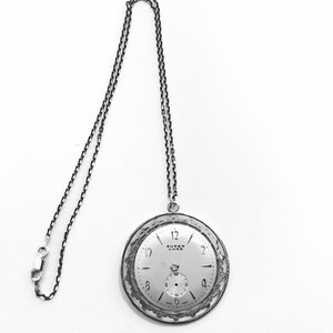 May include: A silver-toned pocket watch pendant with a matching chain. The watch face is light-colored with black numerals and hands. The words "SUPER LUXE" are printed on the watch face. The chain is attached to the top of the watch.