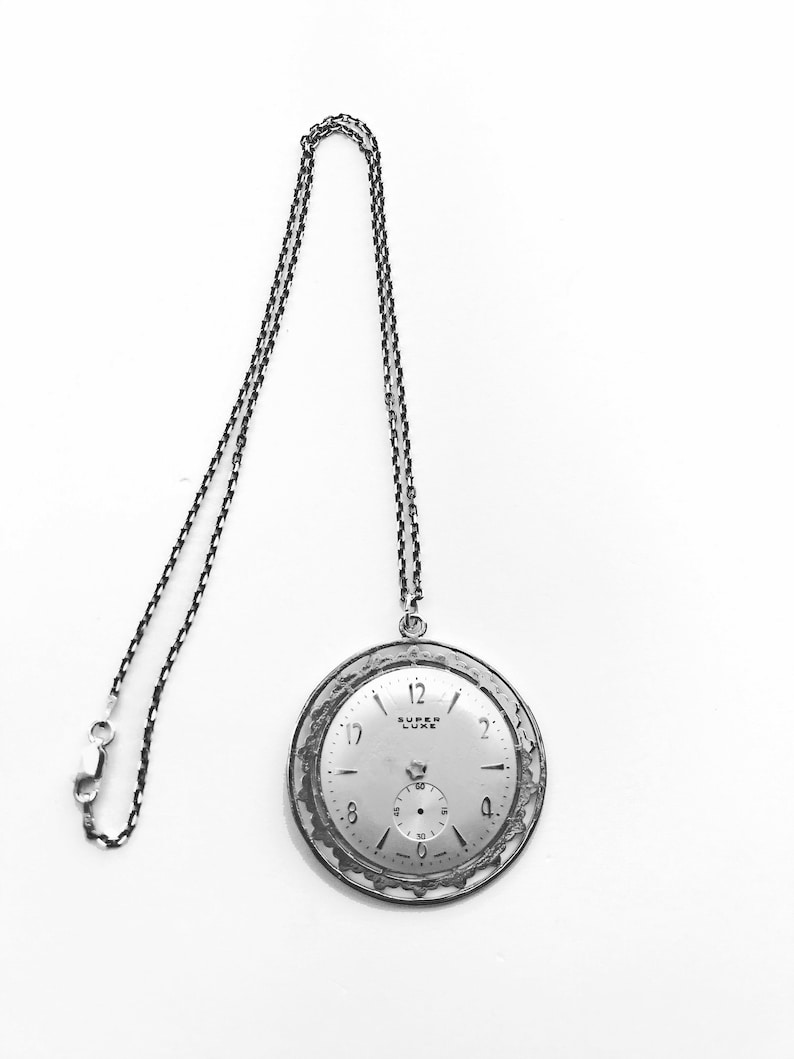 May include: A silver-toned pocket watch pendant with a matching chain. The watch face is light-colored with black numerals and hands. The words "SUPER LUXE" are printed on the watch face. The chain is attached to the top of the watch.