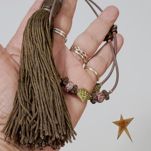 May include: Olive green tassel with a decorative beaded accent. The tassel is attached to a brown cord with decorative beads. The beads are in shades of green, brown, and pink. The tassel is held in a hand.