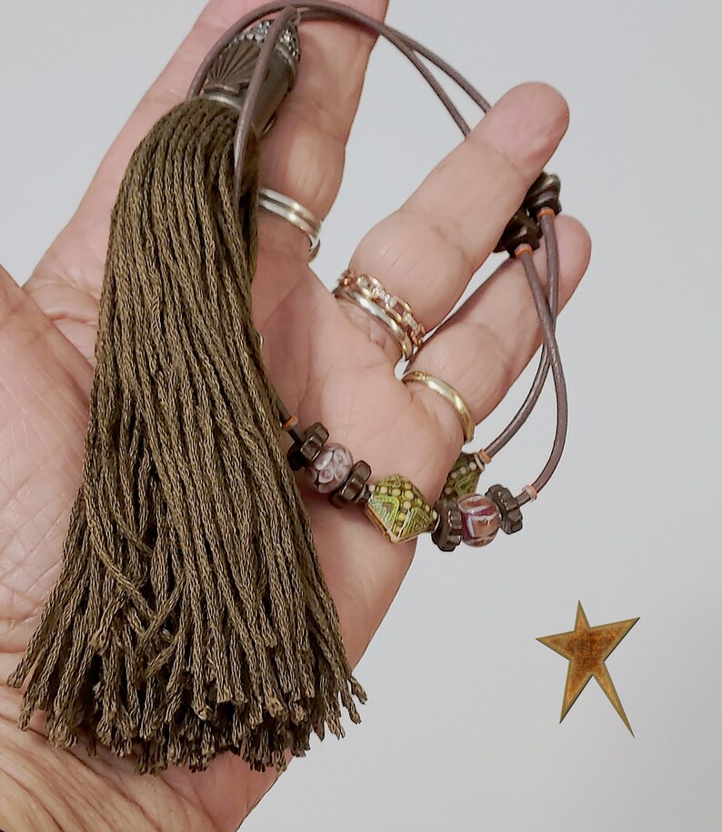 May include: Olive green tassel with a decorative beaded accent. The tassel is attached to a brown cord with decorative beads. The beads are in shades of green, brown, and pink. The tassel is held in a hand.