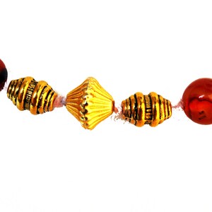 May include: A close-up of a necklace featuring alternating red and gold beads. The red beads are round and marbled, while the gold beads have a textured, metallic appearance. The necklace is strung on a thin, light-colored cord.