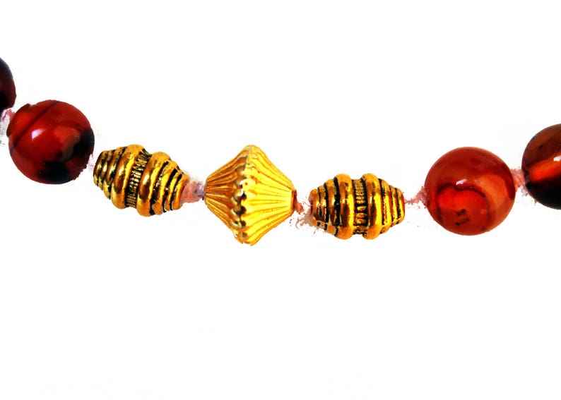 May include: A close-up of a necklace featuring alternating red and gold beads. The red beads are round and marbled, while the gold beads have a textured, metallic appearance. The necklace is strung on a thin, light-colored cord.