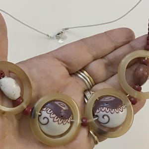 May include: A necklace featuring three large translucent beige circular rings, interspersed with smaller dark red cylindrical beads and two white ceramic beads decorated with brown and black swirling patterns and scalloped line details.