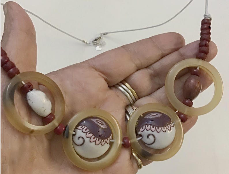 May include: A necklace featuring three large translucent beige circular rings, interspersed with smaller dark red cylindrical beads and two white ceramic beads decorated with brown and black swirling patterns and scalloped line details.