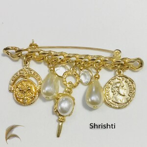 May include: Gold-toned brooch with dangling charms. The brooch features a safety pin design with a chain-like bar. Charms include faux pearls, clear crystals, and coin-shaped pendants. The brooch is set against a white background.