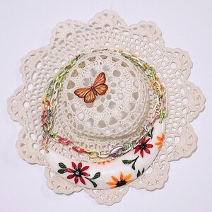 May include: A white crescent-shaped necklace with red and orange floral designs, layered with a colorful chain necklace. A small butterfly sits atop a crocheted sphere, all on a cream-colored doily.