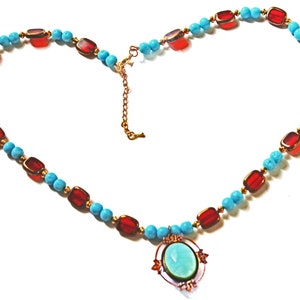May include: A necklace with a heart-shaped design. The necklace features turquoise beads, red rectangular stones, and gold-colored accents. A pendant with a light blue oval stone and gold-colored frame hangs at the center.