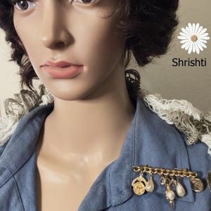 May include: A gold-toned brooch with dangling charms, including pearls and coins, is pinned to a blue denim shirt. The brooch is a decorative accessory. The image also includes the text "Shrishti".