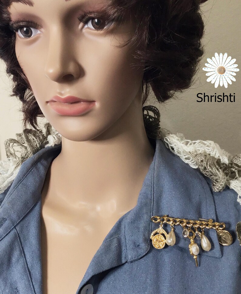 May include: A gold-toned brooch with dangling charms, including pearls and coins, is pinned to a blue denim shirt. The brooch is a decorative accessory. The image also includes the text "Shrishti".