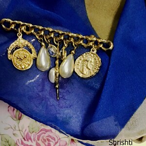 May include: A gold-toned brooch with a chain design, adorned with dangling charms including faux pearls and coin-shaped pendants. The brooch is set against a backdrop of blue and floral fabric.