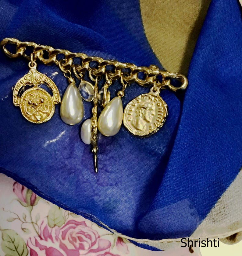 May include: A gold-toned brooch with a chain design, adorned with dangling charms including faux pearls and coin-shaped pendants. The brooch is set against a backdrop of blue and floral fabric.