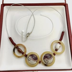 May include: A necklace featuring a silver-toned wire chain with small dark red beads, two large translucent beige rings, and two central circular pendants. The pendants are white with dark purple patterns and decorative swirls, accented by additional beads.