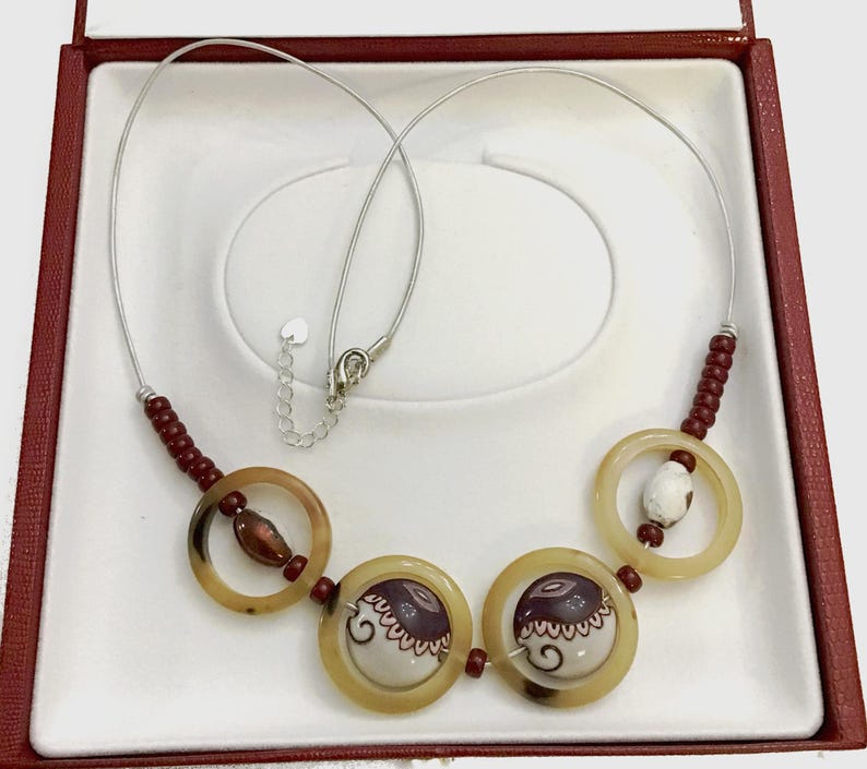 May include: A necklace featuring a silver-toned wire chain with small dark red beads, two large translucent beige rings, and two central circular pendants. The pendants are white with dark purple patterns and decorative swirls, accented by additional beads.
