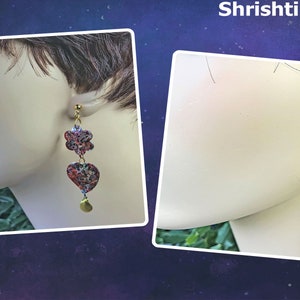 May include: A pair of dangle earrings with a floral and heart-shaped design, and a rectangular and circular design. The earrings are made of a colorful material with red, blue, and gold accents. The earrings are attached to a gold-colored post and have a gold-colored charm at the bottom. The word "Shrishti" is at the top.