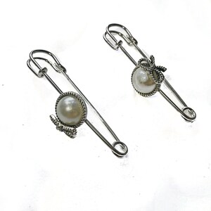 May include: Two silver-toned safety pins with pearl accents. Each pin features a large, round, off-white pearl set in a decorative frame. One pin has a small bow, the other has a small butterfly. The pins are on a white background.