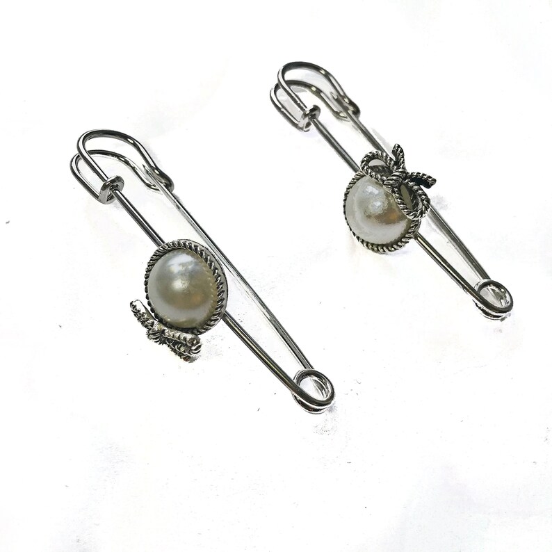 May include: Two silver-toned safety pins with pearl accents. Each pin features a large, round, off-white pearl set in a decorative frame. One pin has a small bow, the other has a small butterfly. The pins are on a white background.