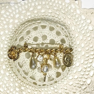May include: Gold-toned brooch with a safety pin backing. The brooch features a chain design with dangling charms, including faux pearls, clear crystals, and coin-shaped pendants. The brooch is set against a white, lacy background.