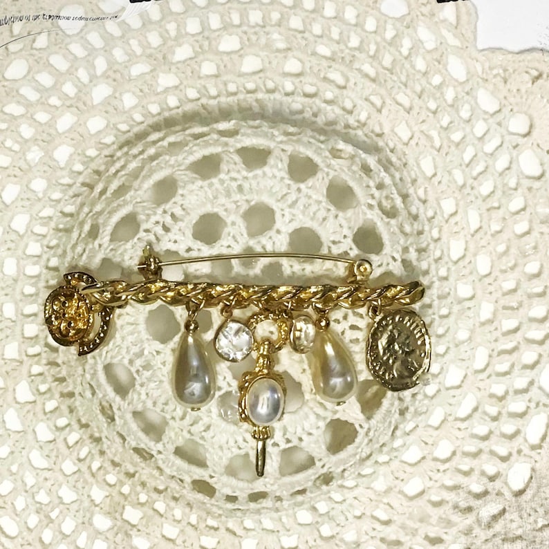 May include: Gold-toned brooch with a safety pin backing. The brooch features a chain design with dangling charms, including faux pearls, clear crystals, and coin-shaped pendants. The brooch is set against a white, lacy background.