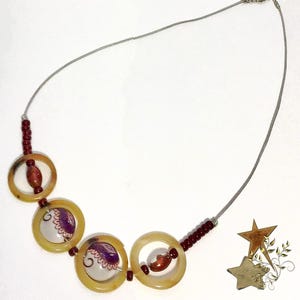 May include: A necklace featuring a thin silver-toned chain, four translucent amber-colored circular rings, and small dark red beads. Two of the rings contain white circular inserts with purple and pink floral patterns, while two contain red beads.