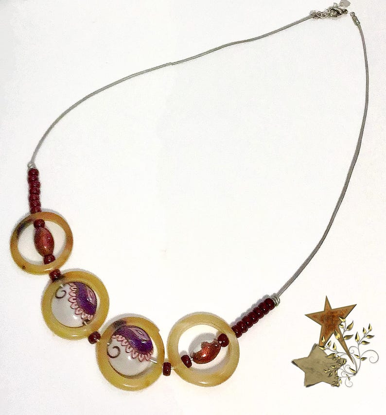 May include: A necklace featuring a thin silver-toned chain, four translucent amber-colored circular rings, and small dark red beads. Two of the rings contain white circular inserts with purple and pink floral patterns, while two contain red beads.