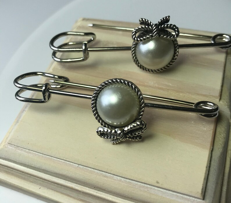 May include: Two silver-toned safety pins with pearl-like centers and decorative bow accents. The pins are displayed on a light-colored surface, showcasing their elegant design. The pins are likely used for fashion or craft projects.