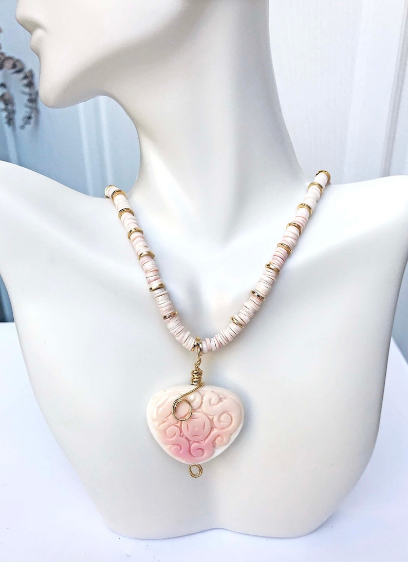 May include: A necklace with a heart-shaped pendant. The pendant is a light pink color with a carved design and is suspended from a necklace of white and gold beads. The necklace is displayed on a white mannequin.