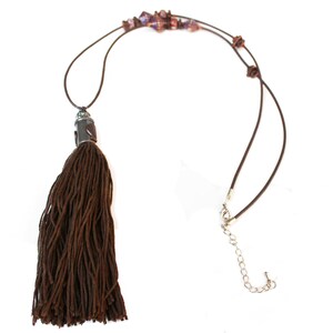 May include: A brown tassel necklace with a dark brown cord. The necklace features a decorative metal piece at the top of the tassel and small, colorful beads along the cord. The necklace has a silver-colored clasp and extender chain.