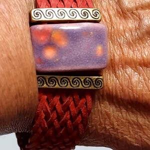 May include: Red braided leather bracelet with a rectangular purple and orange ceramic centerpiece. The bracelet features gold-colored metal accents with a spiral design. A close-up shot highlights the texture and color of the jewelry.