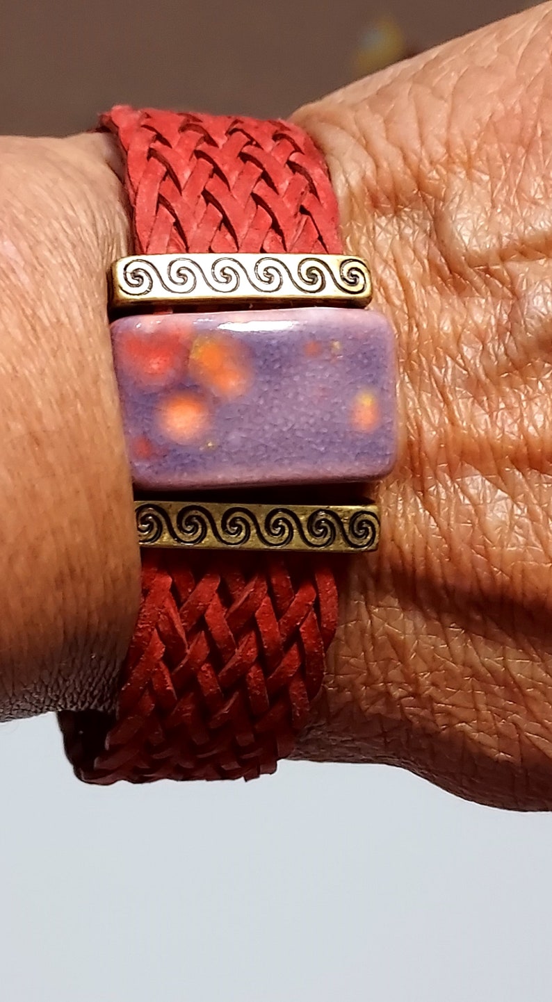 May include: Red braided leather bracelet with a rectangular purple and orange ceramic centerpiece. The bracelet features gold-colored metal accents with a spiral design. A close-up shot highlights the texture and color of the jewelry.