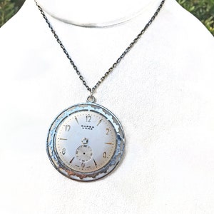 May include: A silver-toned pendant necklace featuring a vintage watch face. The watch face is white with black numerals and hands, and the text "SUPER LUXE" is visible. The pendant hangs from a dark chain.