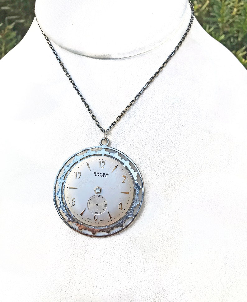 May include: A silver-toned pendant necklace featuring a vintage watch face. The watch face is white with black numerals and hands, and the text "SUPER LUXE" is visible. The pendant hangs from a dark chain.