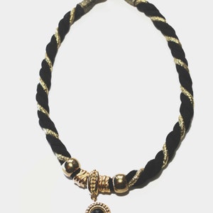May include: A black and gold rope necklace with a gold-toned clasp and accents. The necklace features a gold pendant with a black stone center. The rope is twisted with gold metallic thread.