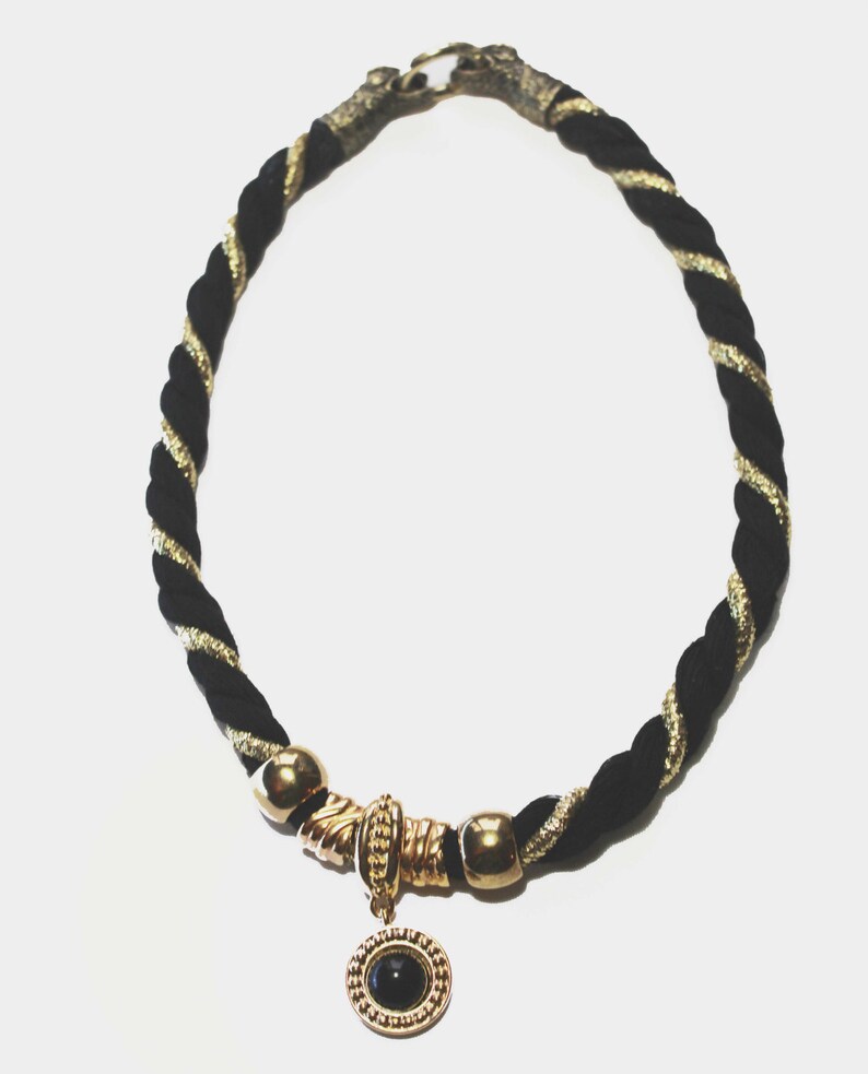 May include: A black and gold rope necklace with a gold-toned clasp and accents. The necklace features a gold pendant with a black stone center. The rope is twisted with gold metallic thread.