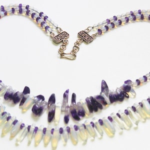 May include: A two-strand necklace featuring iridescent, teardrop-shaped beads and small purple beads. The necklace has a silver clasp and is designed with a heart-like shape. The beads are arranged in a symmetrical pattern.