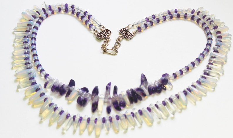 May include: A two-strand necklace featuring iridescent, teardrop-shaped beads and small purple beads. The necklace has a silver clasp and is designed with a heart-like shape. The beads are arranged in a symmetrical pattern.
