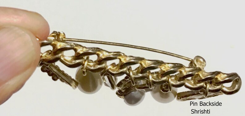 May include: Backside view of a gold-toned brooch with a chain design and dangling faux pearls. The pin has a safety clasp. The text "Pin Backside Shrishti" is visible.