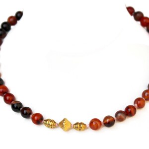 May include: A beaded necklace featuring alternating red and black round beads. The necklace has three gold-colored, decorative beads in the center. The beads are strung on a thin, light-colored string. The necklace is displayed on a white background.