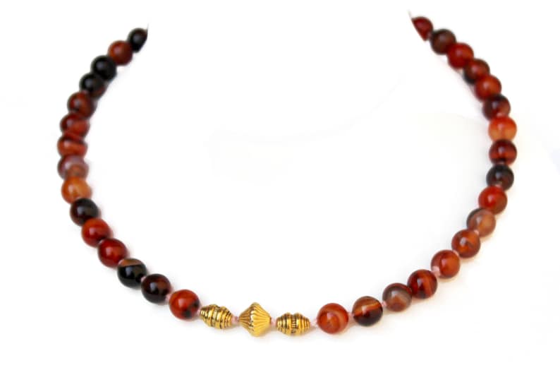 May include: A beaded necklace featuring alternating red and black round beads. The necklace has three gold-colored, decorative beads in the center. The beads are strung on a thin, light-colored string. The necklace is displayed on a white background.