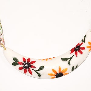May include: A white, curved necklace pendant with a floral design. The pendant features red and orange flowers with green leaves. The chain is made of colorful, painted links. A unique, handcrafted jewelry piece.