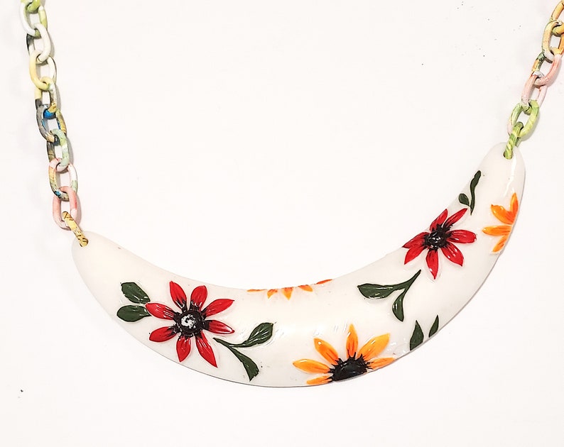 May include: A white, curved necklace pendant with a floral design. The pendant features red and orange flowers with green leaves. The chain is made of colorful, painted links. A unique, handcrafted jewelry piece.