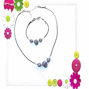 Op de afbeelding: Een zwarte ketting- en armbandset met schedelhangers met rode ogen en zilveren accenten. De sieraden worden gepresenteerd op een witte achtergrond, met decoratieve bloemen en knopen in de hoeken.