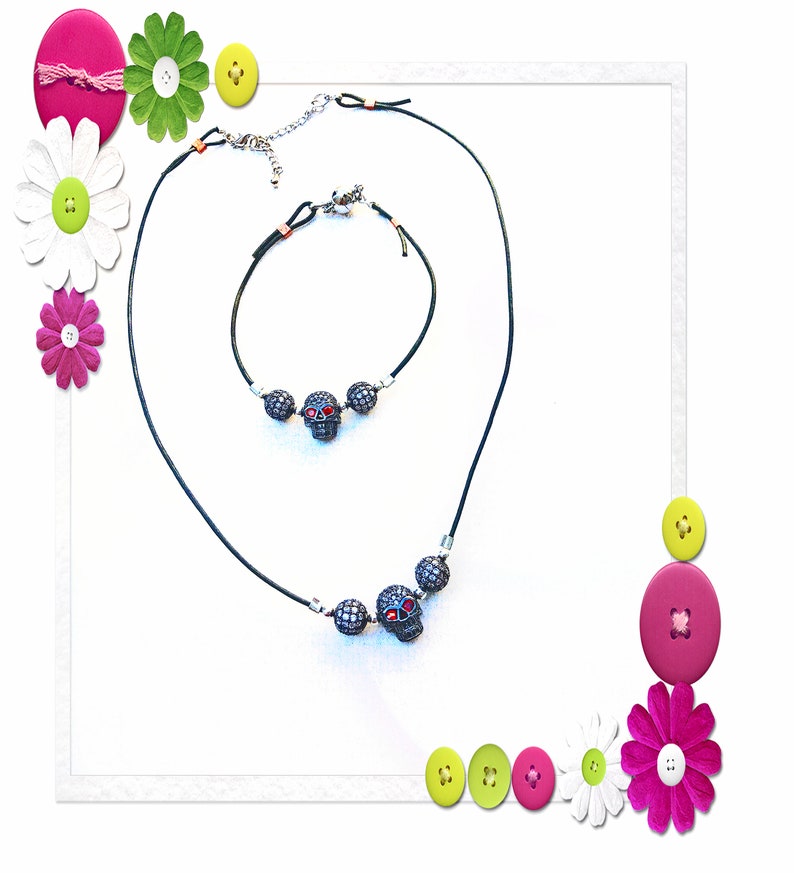 Op de afbeelding: Een zwarte ketting- en armbandset met schedelhangers met rode ogen en zilveren accenten. De sieraden worden gepresenteerd op een witte achtergrond, met decoratieve bloemen en knopen in de hoeken.