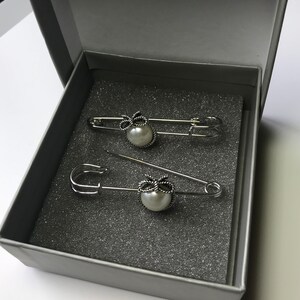 May include: Two silver-toned safety pins with a pearl and bow design, presented in a gray box. The pins are arranged on a gray foam insert. The pearls are white and the bows are detailed.