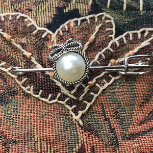 May include: A silver-toned brooch with a large, round, faux pearl center. The pearl is surrounded by a twisted rope design and topped with a bow. The brooch has a pin closure and is set against a patterned fabric.